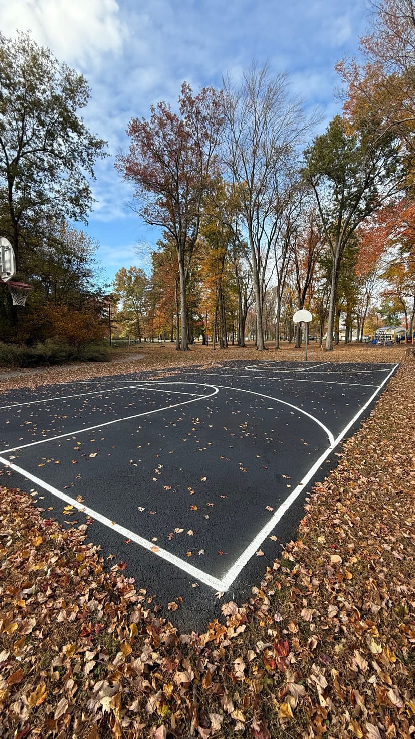 Empty basketball court surrounded by autumn leaves and colorful trees under a blue sky.